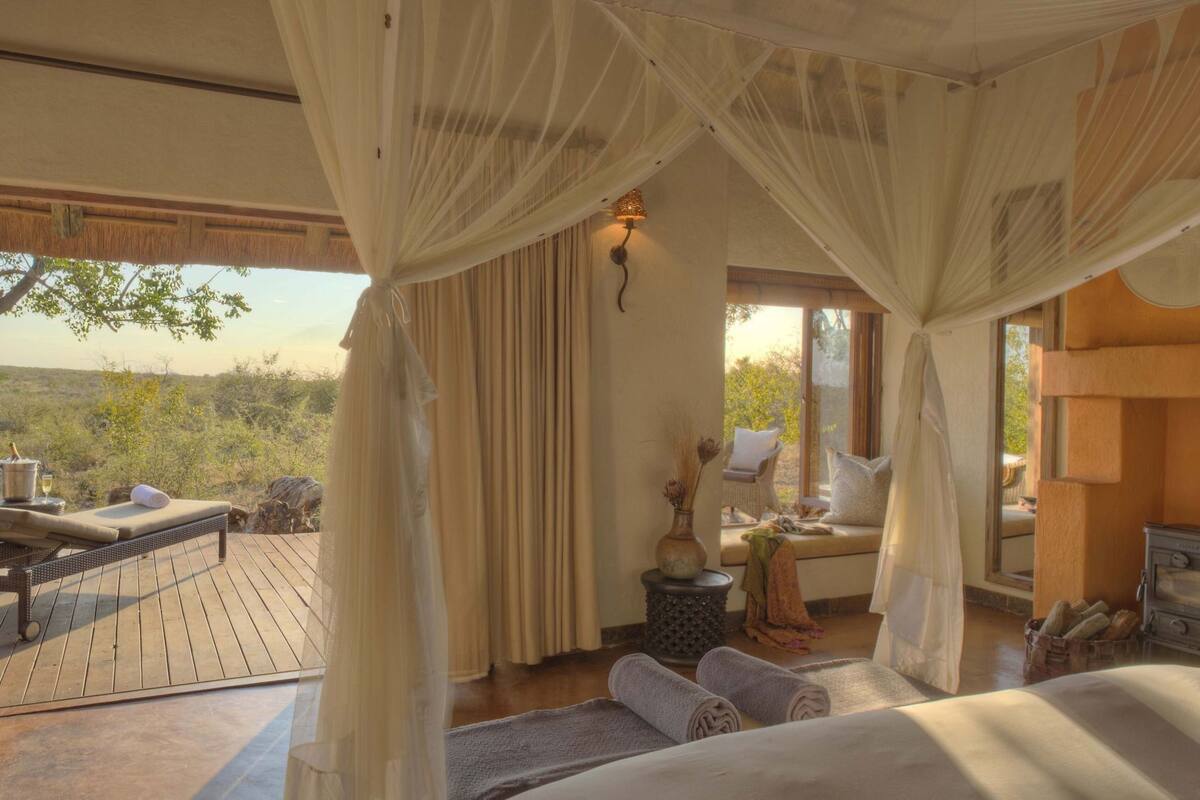 A room with a bed, a small table with a lamp, a window with curtains, and a view of the outdoors.