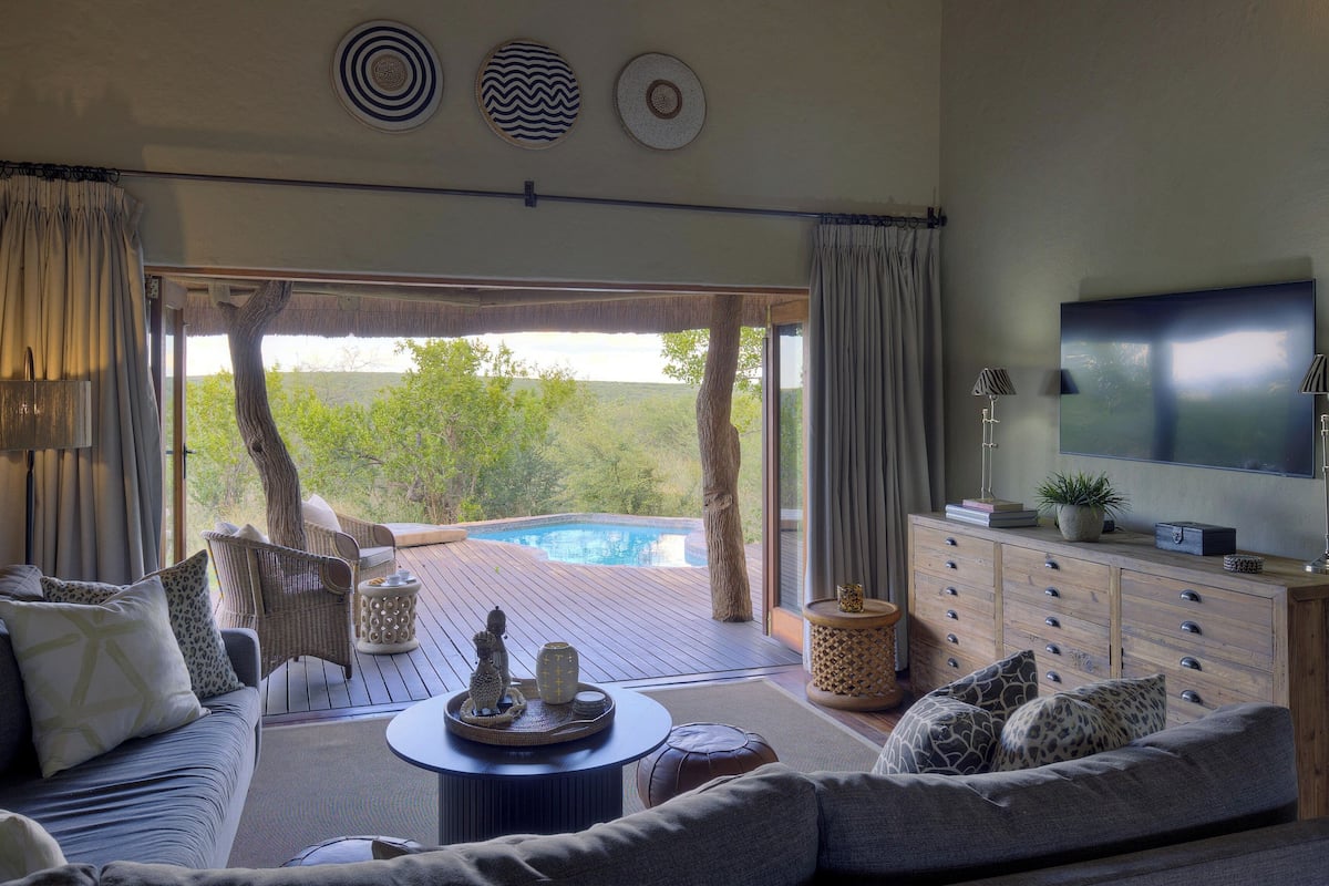 A spacious living room with a large sofa, a coffee table, and a TV. There is a view of a pool and greenery outside.