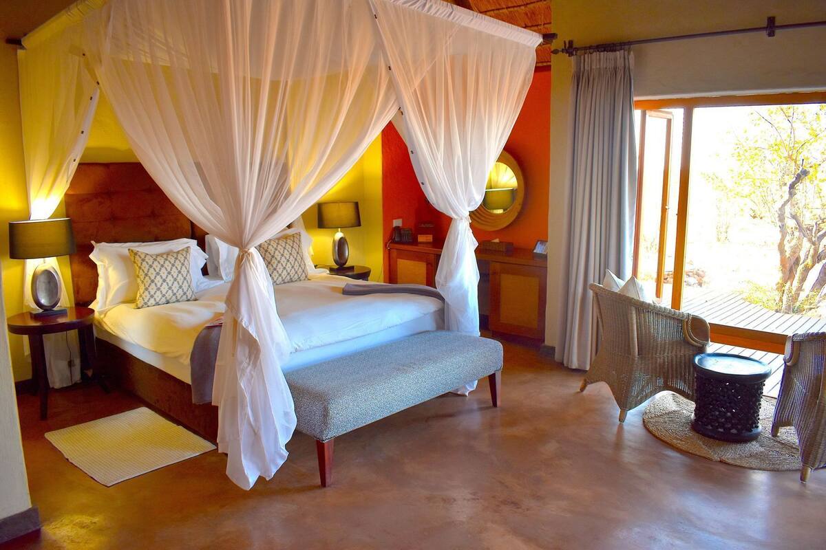 A room with a bed, bedside lamps, a bench, a table with chairs, and a window.