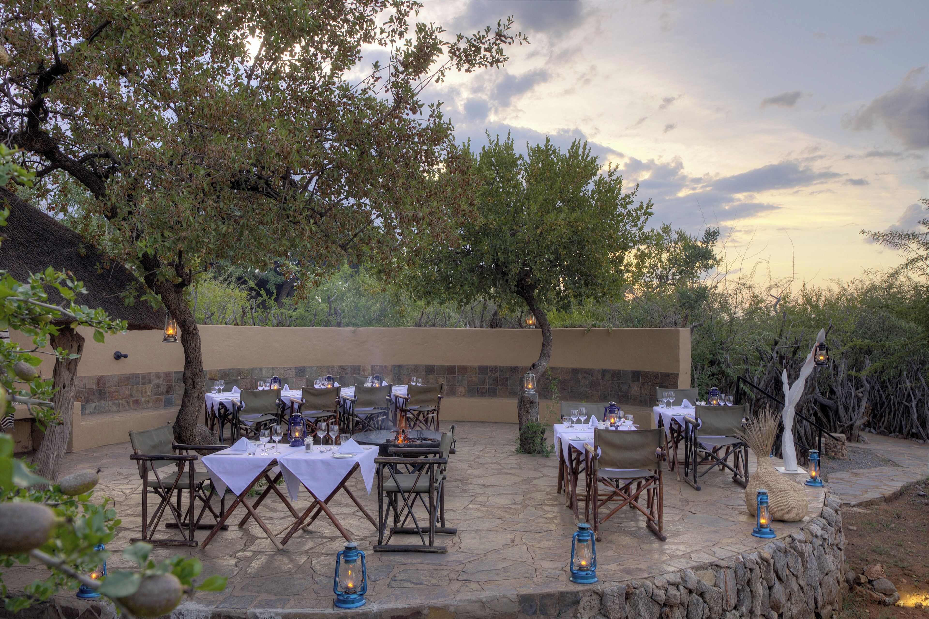 An outdoor dining area with tables set for a meal, surrounded by stone walls and trees.