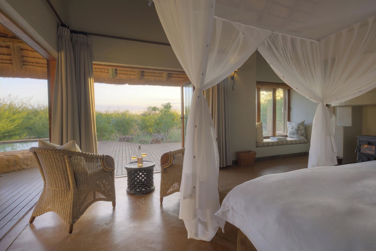 A bedroom with a large bed, a wicker chair, a small table with candles, and a view of greenery outside.