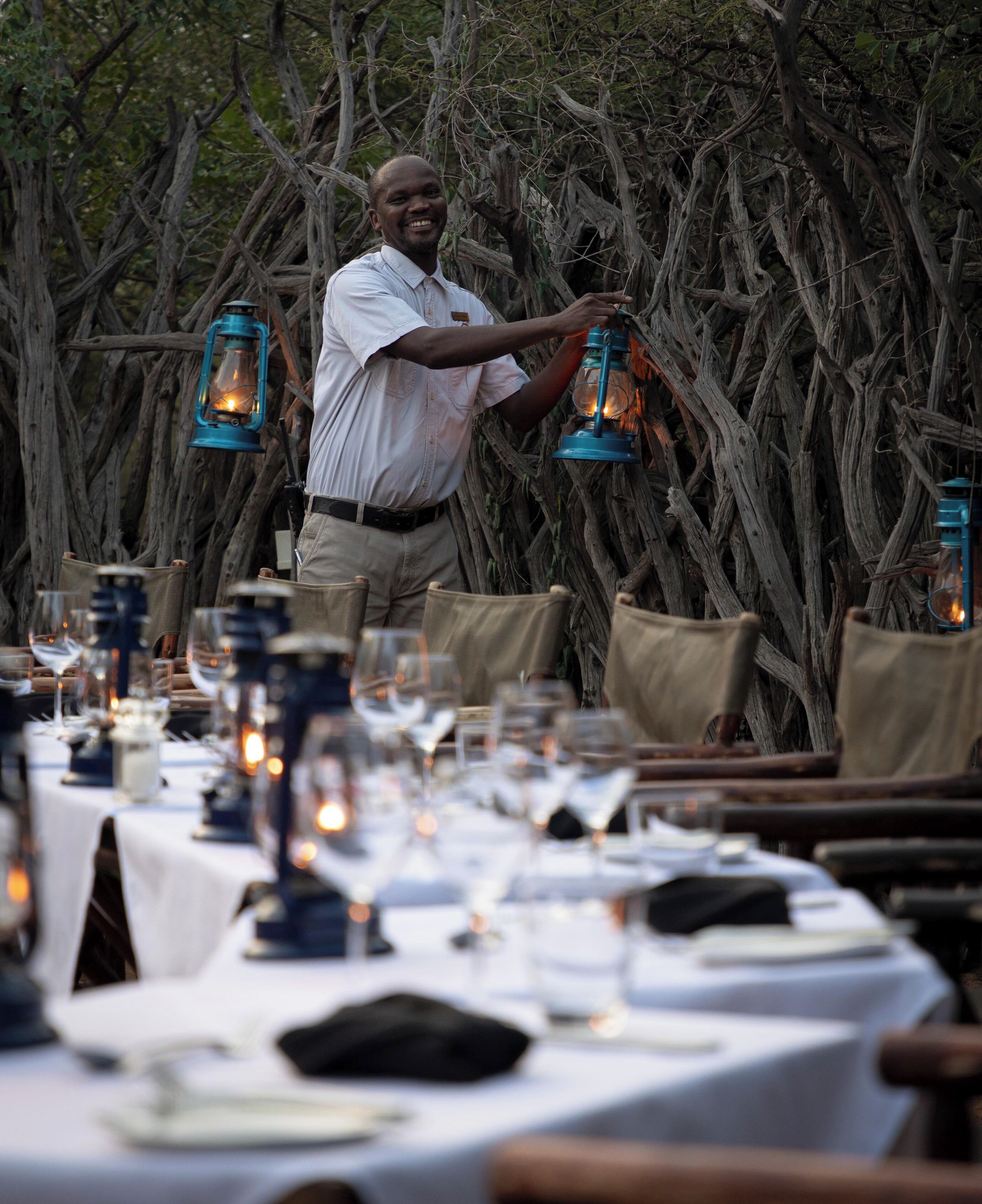 A man arranging lanterns on a table set outdoors with a natural backdrop of twisted tree branches.