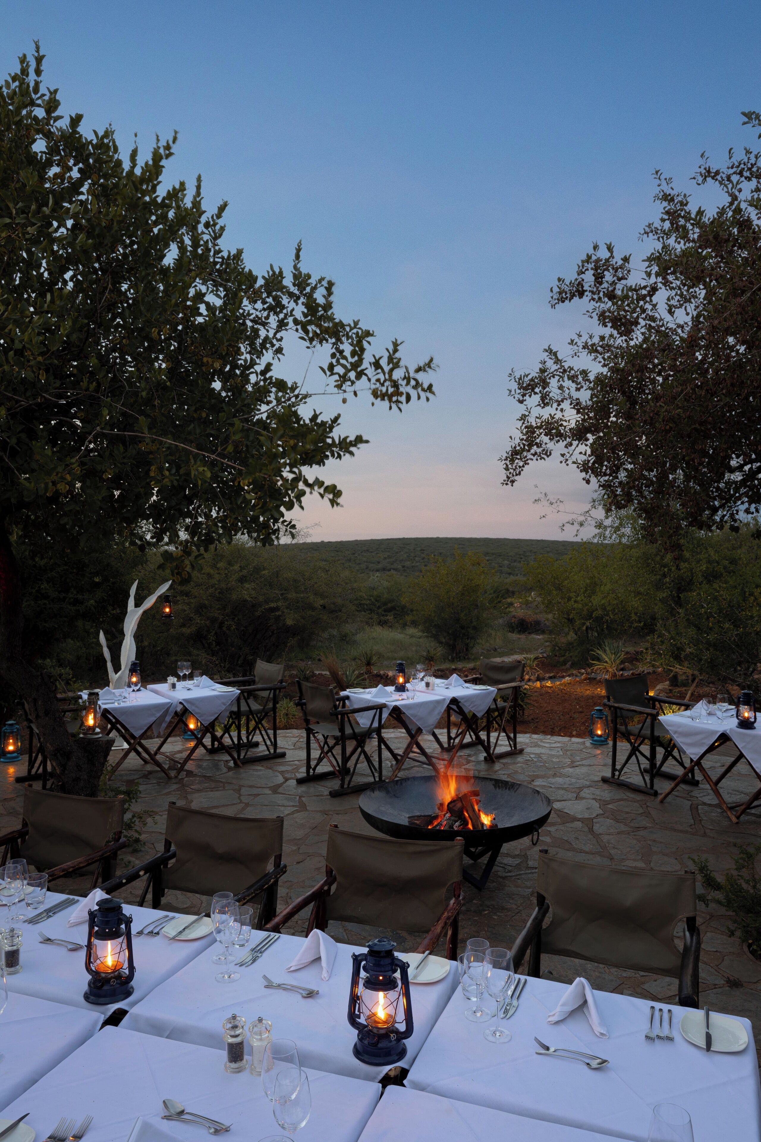 An outdoor dining area with tables set for two, surrounded by chairs, and a fire pit in the center.