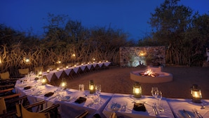 Un área de comedor al aire libre con mesas dispuestas para una comida, rodeada de un muro de piedra y vegetación.