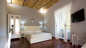 A bedroom with a wooden floor, a bed with a floral bedspread, a nightstand, a dresser, a television on the wall, and a window with curtains. - Residenza Sistina (Rome)