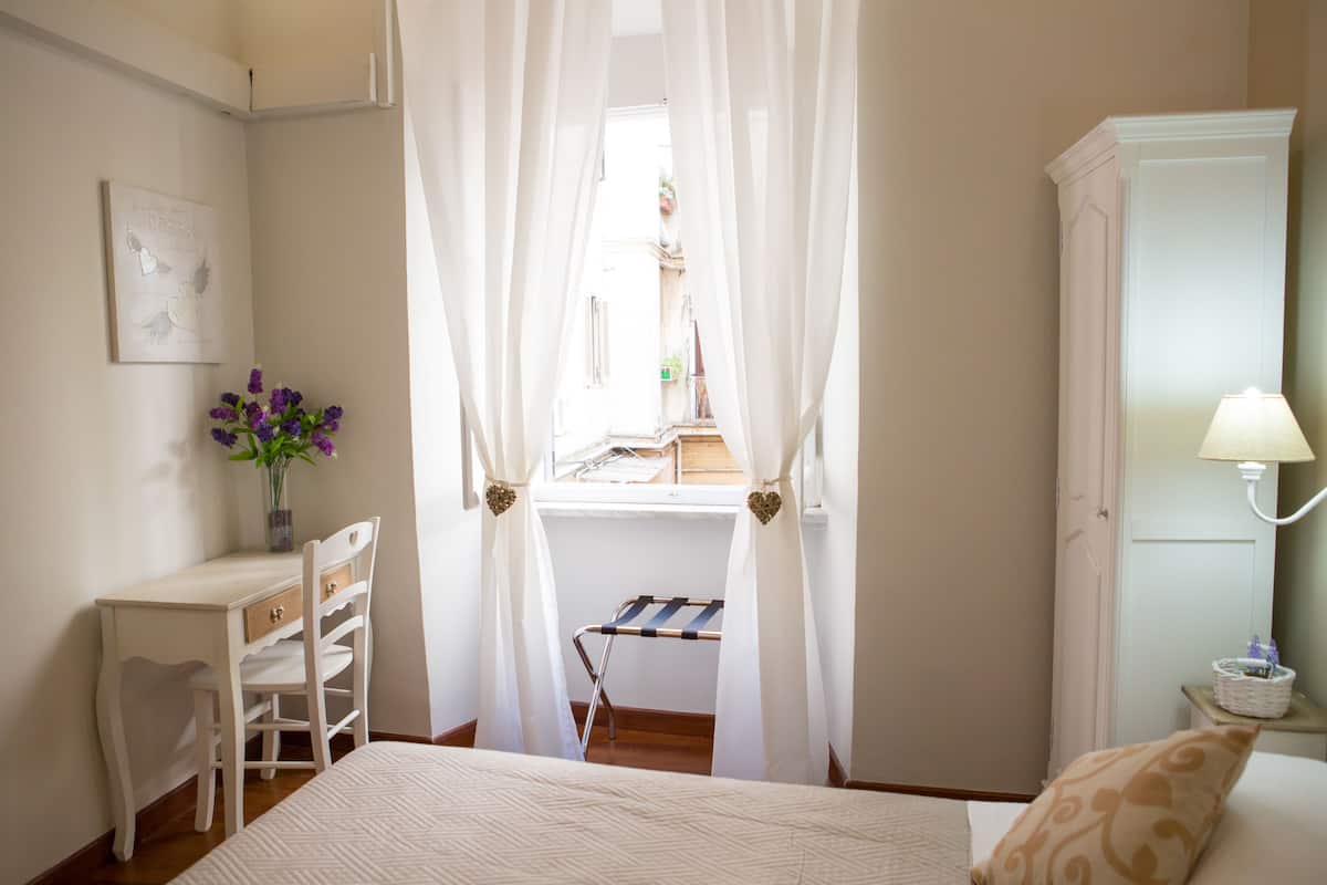 A bedroom with a bed, a desk with a chair, a window with curtains, a lamp, and a wardrobe.