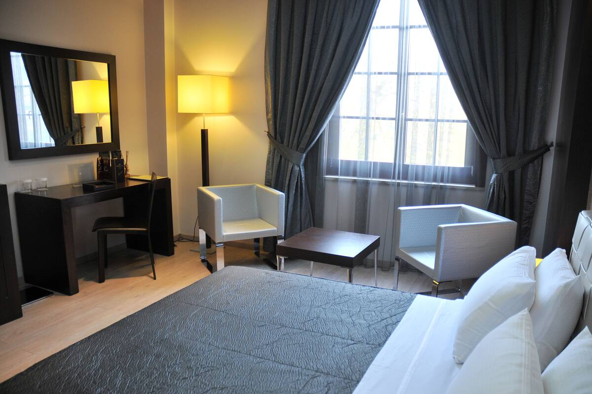 A hotel room with a bed, two armchairs, a desk, a lamp, and a large window with curtains.