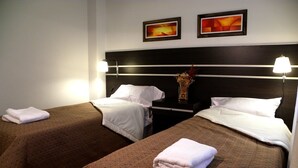 A hotel room with two single beds, each with a brown bedspread and white pillows. There are two bedside tables with lamps, and a small vase with dried flowers on one table. - Santiago Building (Córdoba)