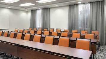 A conference room with a long table and rows of chairs, large windows with curtains, and a carpeted floor.