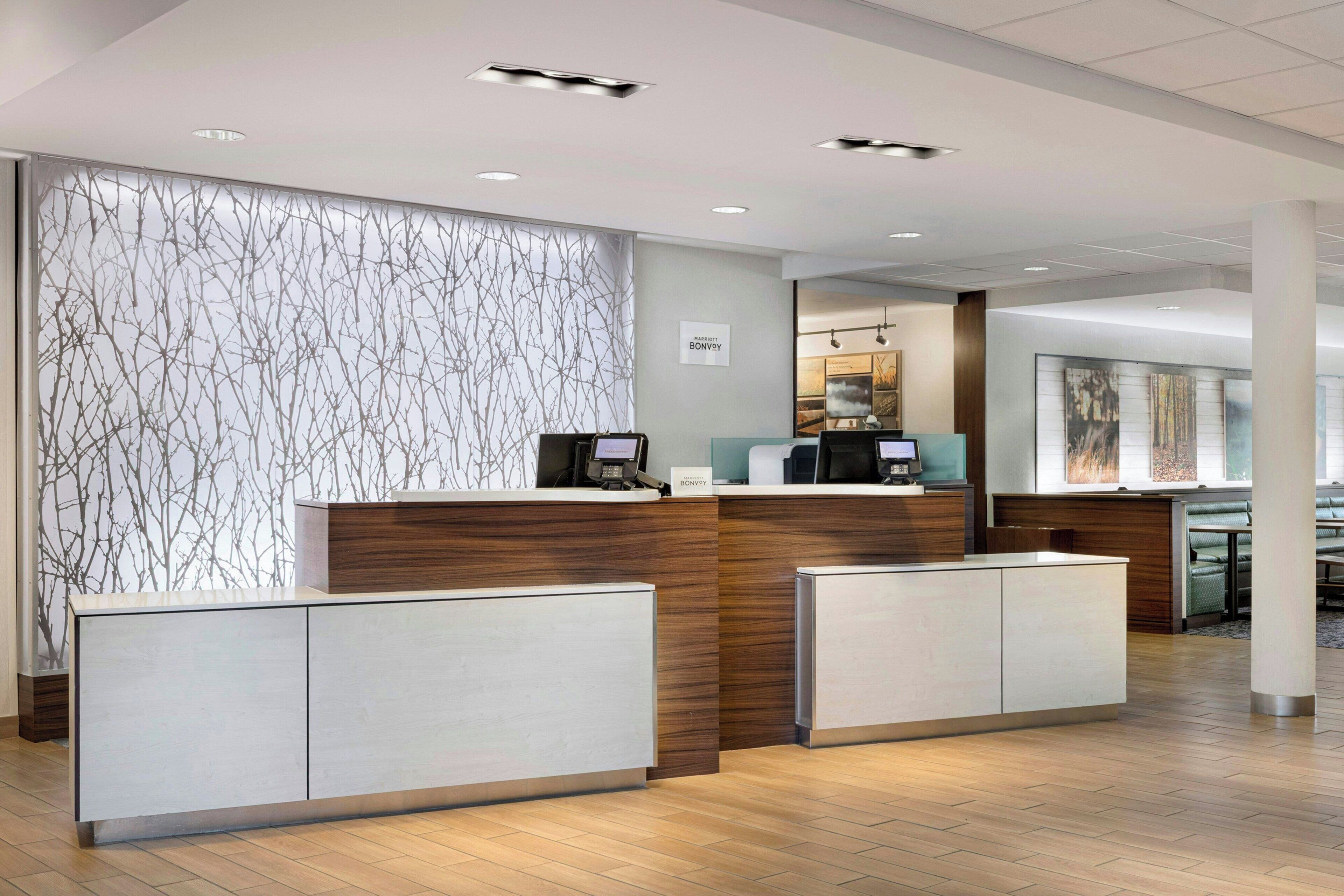 A hotel reception area with a modern design, featuring a wooden reception desk, a digital screen, and a seating area with a view of the cityscape.