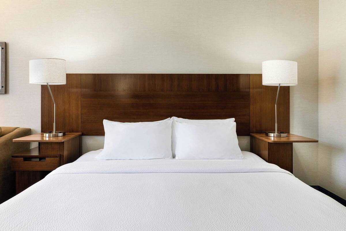 Una habitación de hotel con cabecera de madera, dos mesitas de noche con lámparas, una cama con ropa blanca y un sillón marrón.