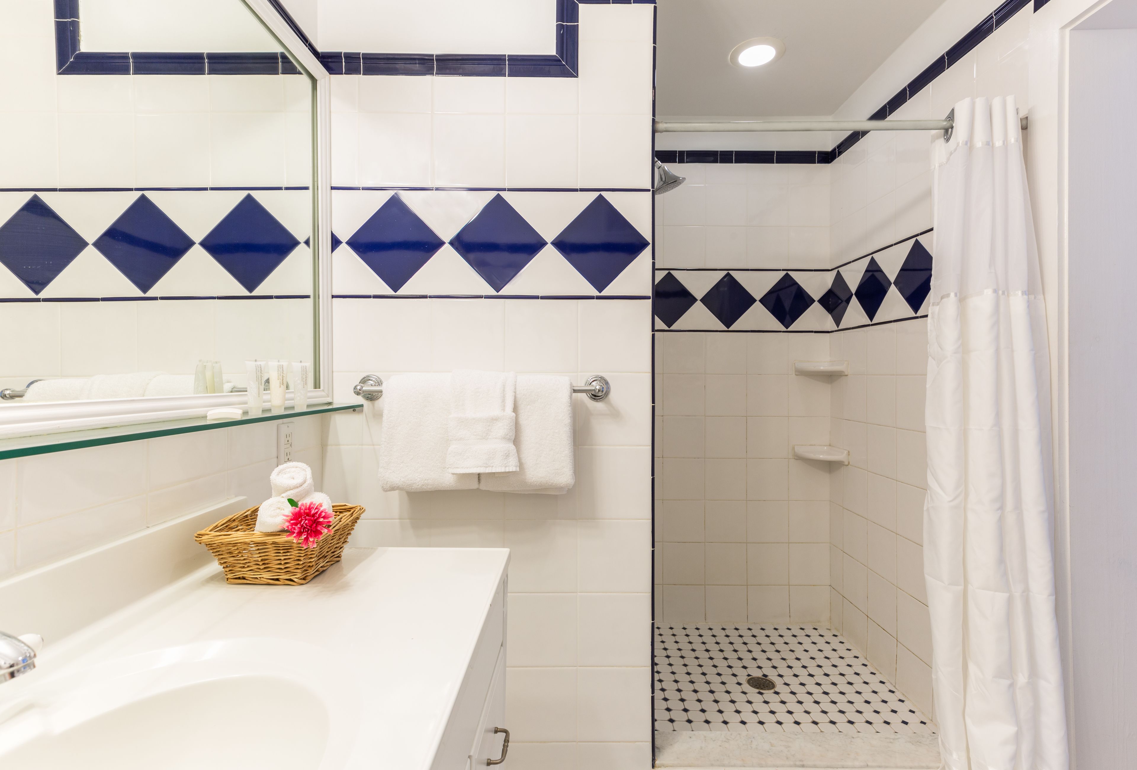 Une salle de bain avec un lavabo blanc, une étagère en verre, une serviette blanche, une douche avec un sol à carreaux noirs et blancs, et des carreaux bleus à motifs losangés sur les murs.