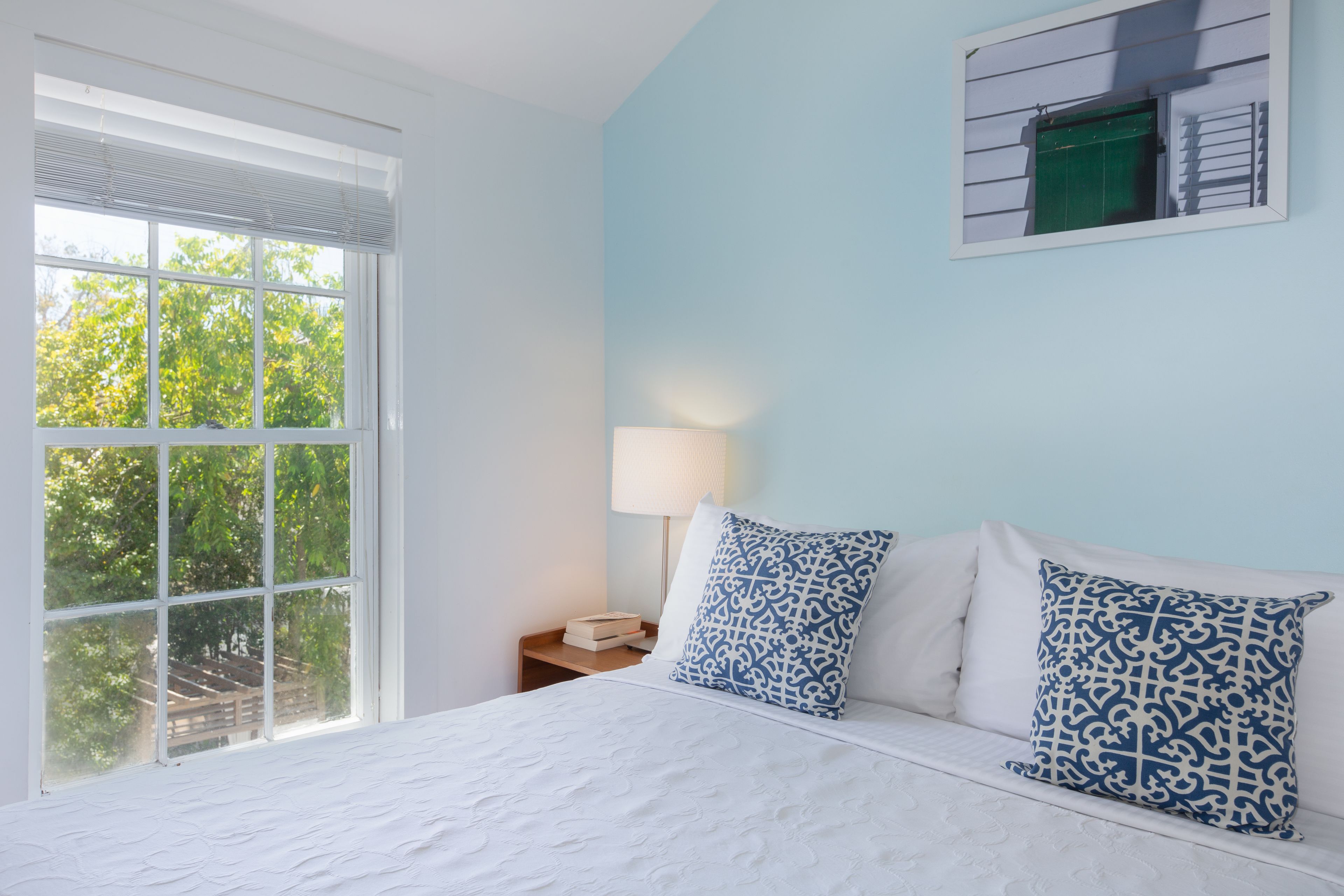 A bedroom with a large window, a bed with white bedding, two patterned pillows, a bedside table with a lamp, and a wall-mounted air conditioning unit.