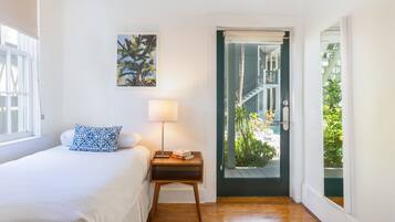 A bedroom with a bed, a nightstand with a lamp, a framed picture on the wall, and a door leading to a balcony with plants.
