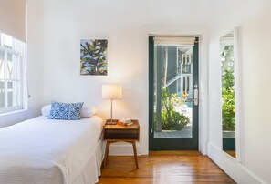 A bedroom with a bed, a nightstand with a lamp, a framed picture on the wall, and a door leading to a balcony with plants.