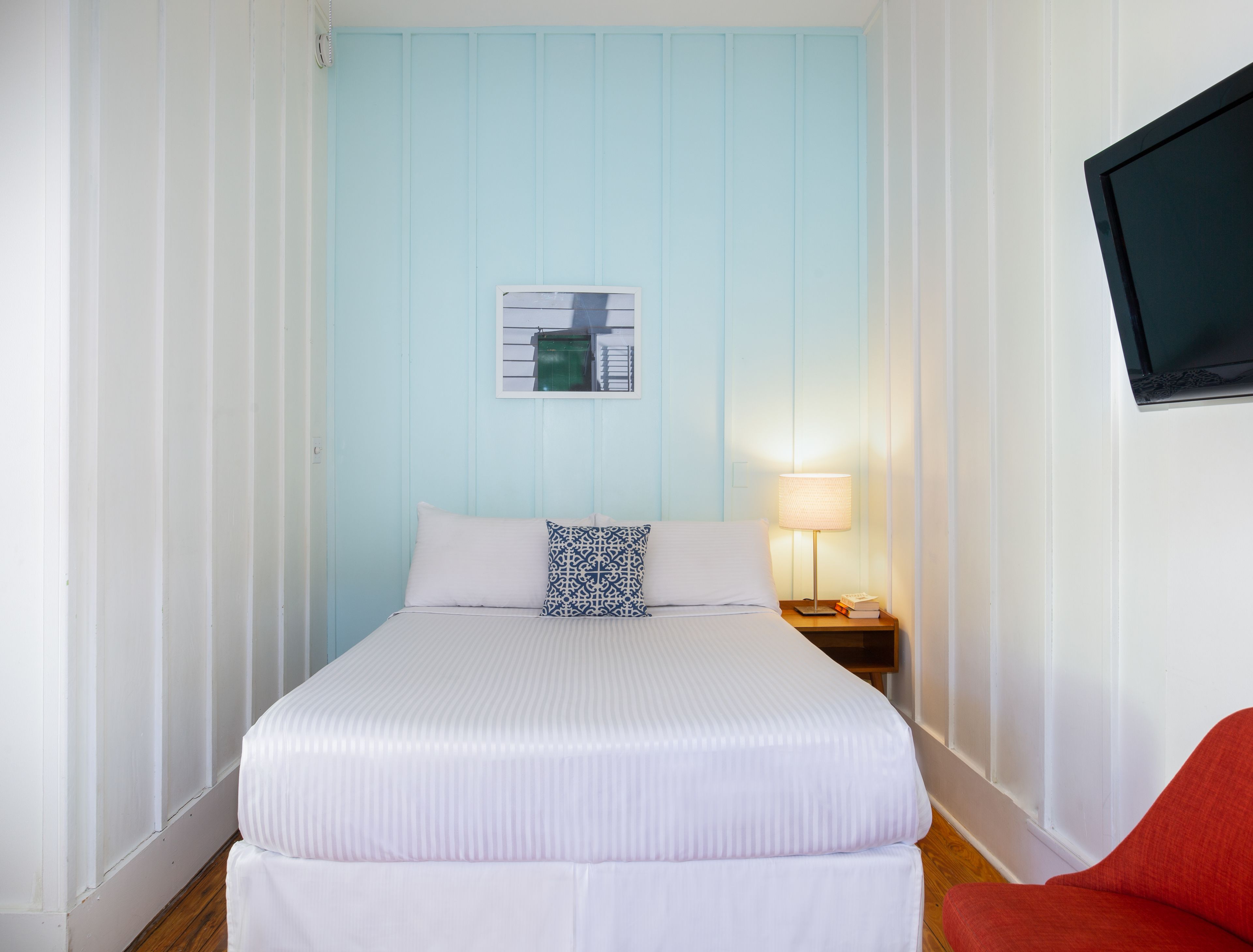 Ein ordentlich bezogenes Bett mit weißer Bettwäsche und einem blau-weißen Kopfkissen, ein Nachttisch mit Lampe und ein roter Sessel in einem Zimmer mit hellblauen Wänden und einem angebrachten Fernseher.