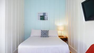 Ein ordentlich bezogenes Bett mit weißer Bettwäsche und einem blau-weißen Kopfkissen, ein Nachttisch mit Lampe und ein roter Sessel in einem Zimmer mit hellblauen Wänden und einem angebrachten Fernseher.