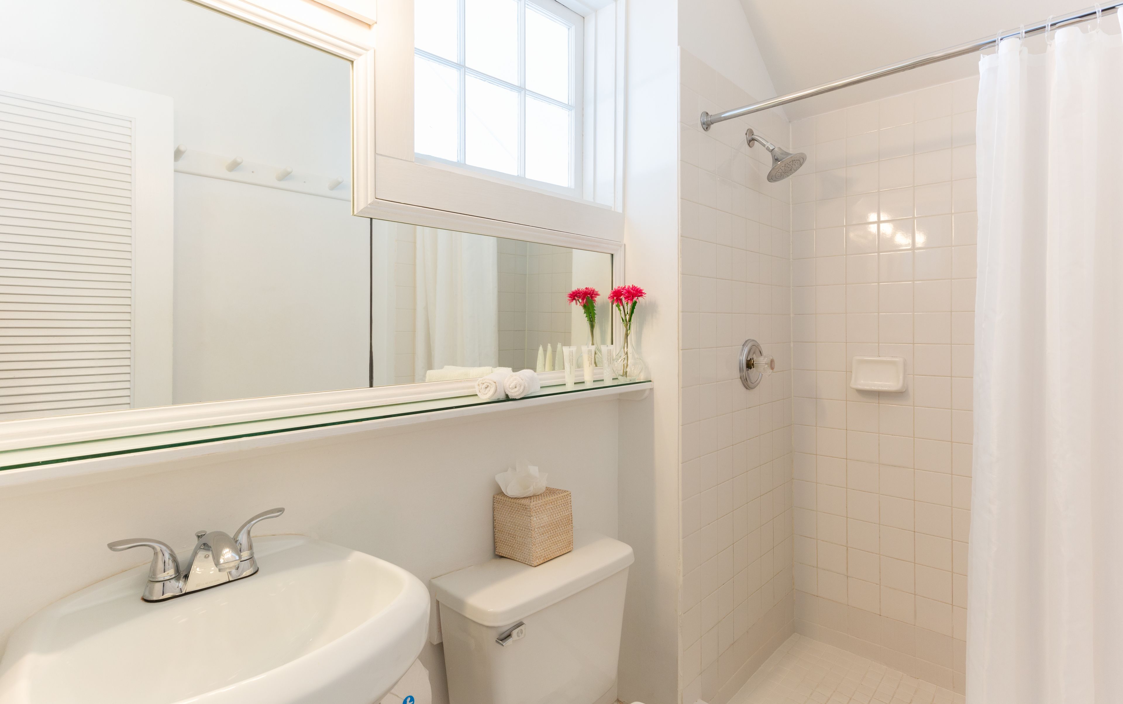 A bathroom with a white sink, a toilet, a glass shelf with toiletries, a shower with a glass door, and a window above the mirror.