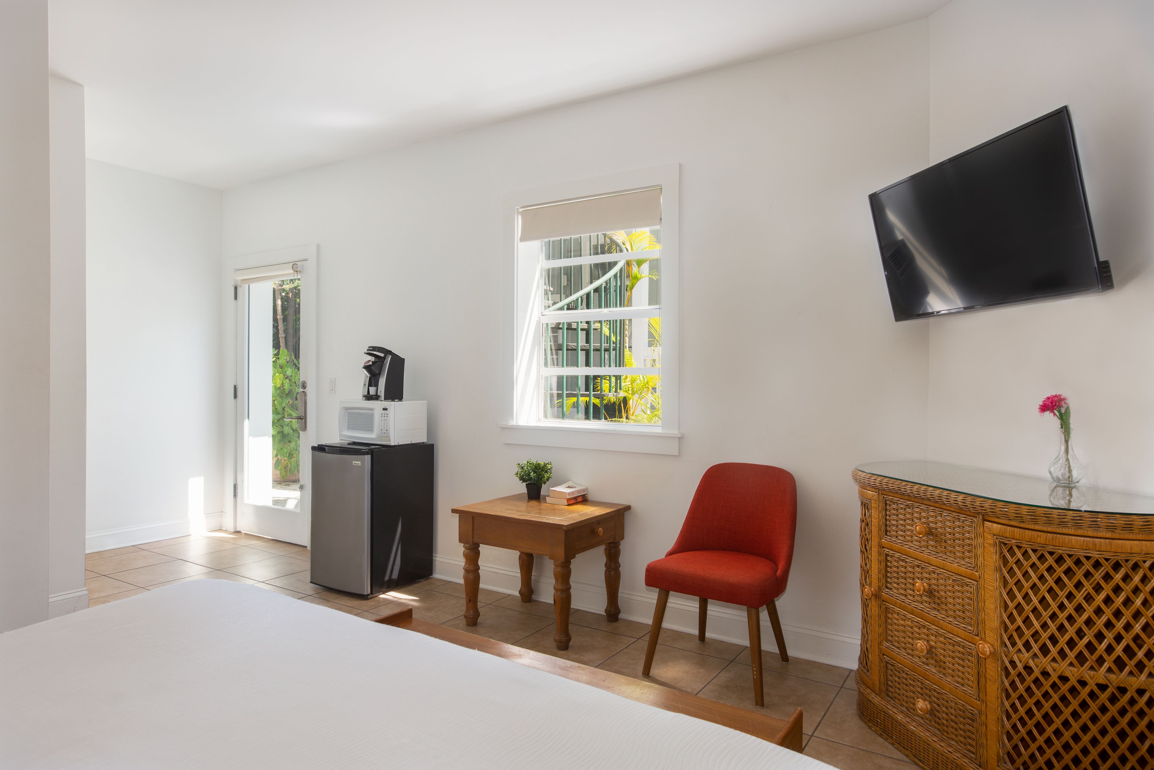 Une pièce avec un lit, une petite table sur laquelle est posée une plante, une chaise rouge, un téléviseur fixé au mur et un réfrigérateur.