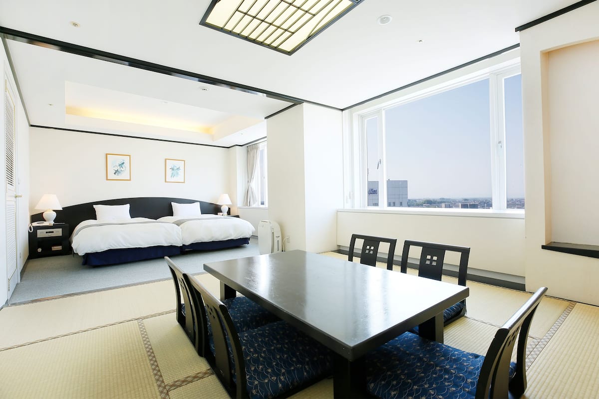 A hotel room with a bed, a dining table, chairs, a view of the city, and a large window.