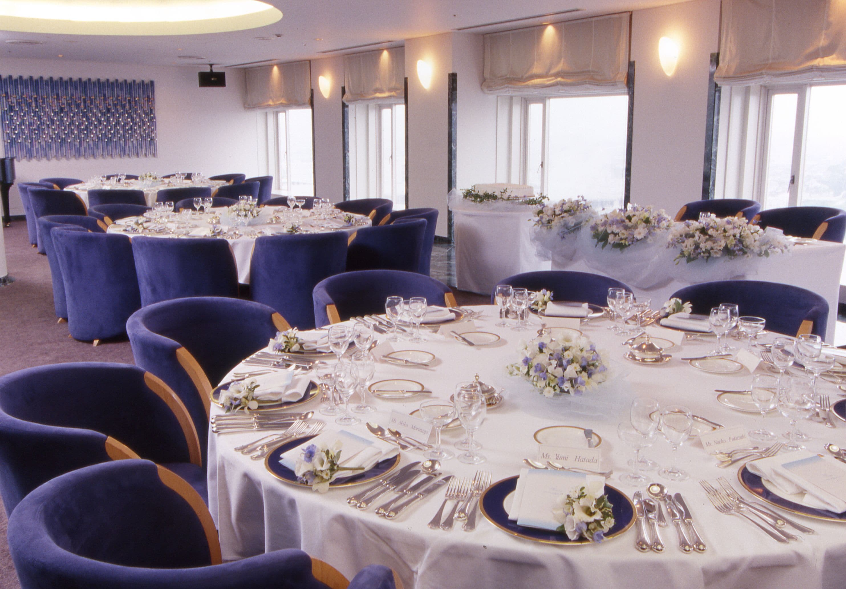 Un salón de banquetes con mesas redondas dispuestas para un evento formal, con manteles blancos, sillas azules y centros de mesa florales.