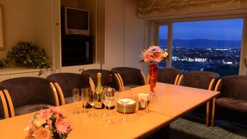 Un coin repas avec une table mise pour le dîner, un téléviseur fixé au mur et une vue sur la ville au crépuscule.