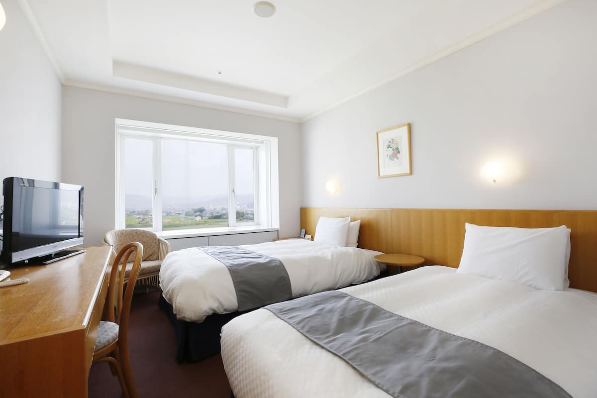 A hotel room with two beds, a desk with a TV, a chair, a window with a view, and a framed picture on the wall.