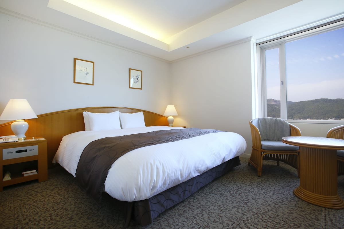 A hotel room with a large bed, two bedside lamps, a chair, a small table, and a window with a view of the hills.
