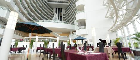 Ein mehrgeschossiges Atrium mit Esstischen, Stühlen und Sonnenschirmen, mit einer zentralen Glasdecke und weißen Trageelementen.