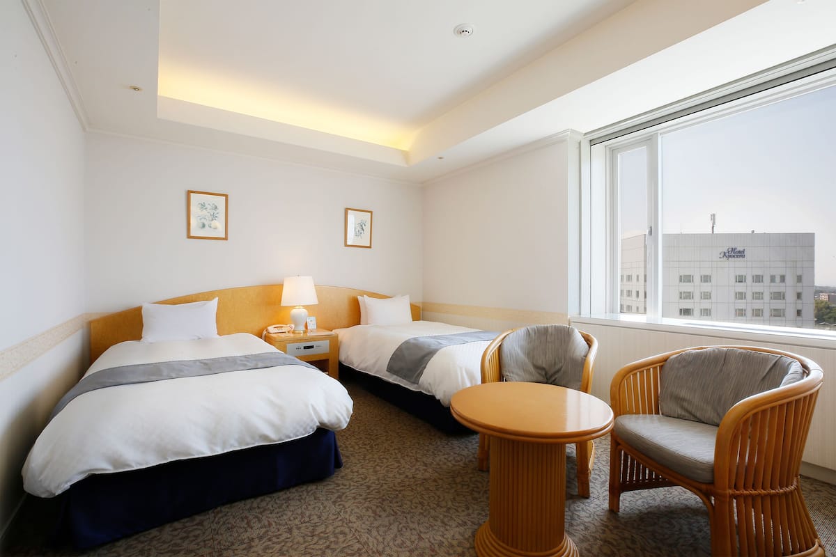 A hotel room with two beds, a chair, a nightstand, and a window with a view of a building.