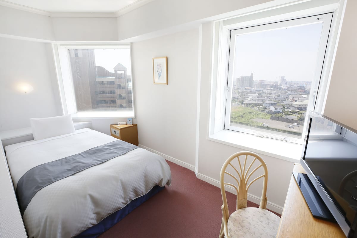 A hotel room with a bed, a chair, a TV, a window with a city view, and a small table.