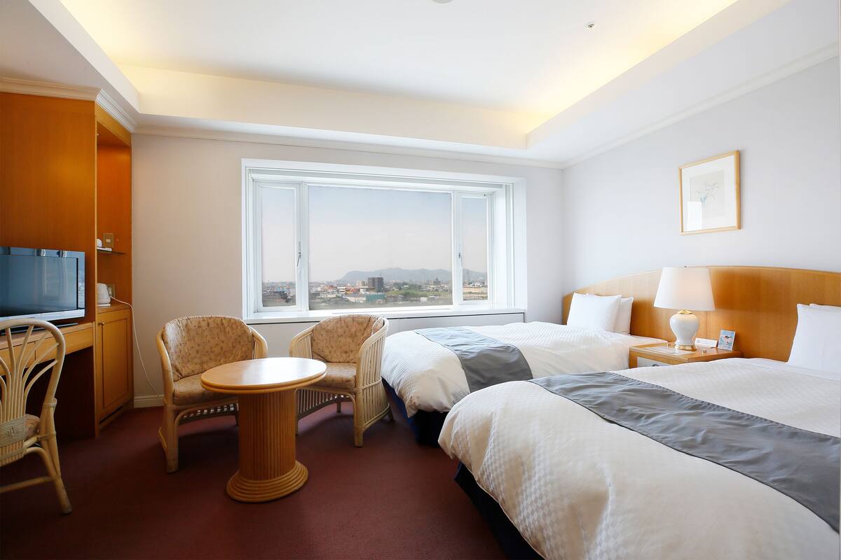 A hotel room with two beds, a desk with a chair, a small table, a TV, and a window with a city view.