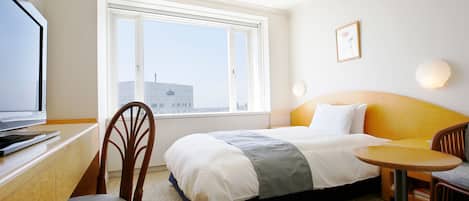 A hotel room with a bed, a desk with a laptop, a chair, a small round table, and a window with a city view.