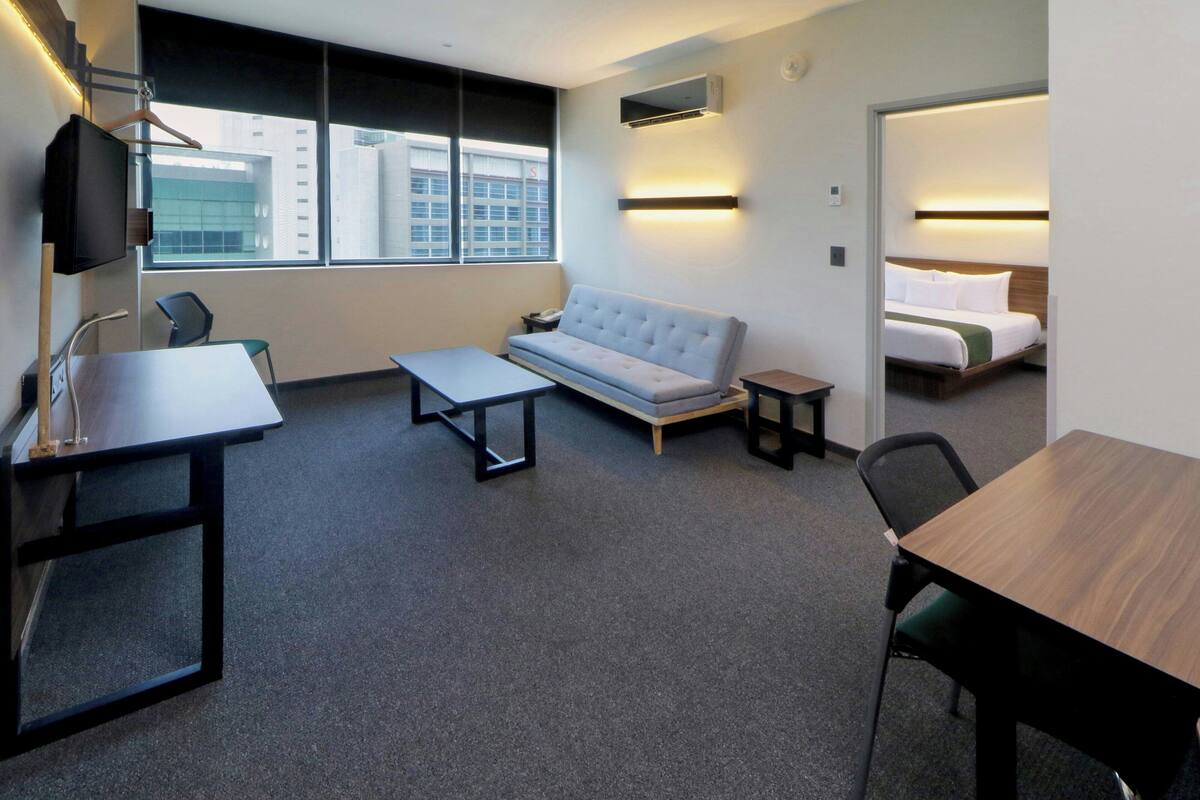 A modern hotel room with a sofa, a desk, a bed, and a view of the cityscape.