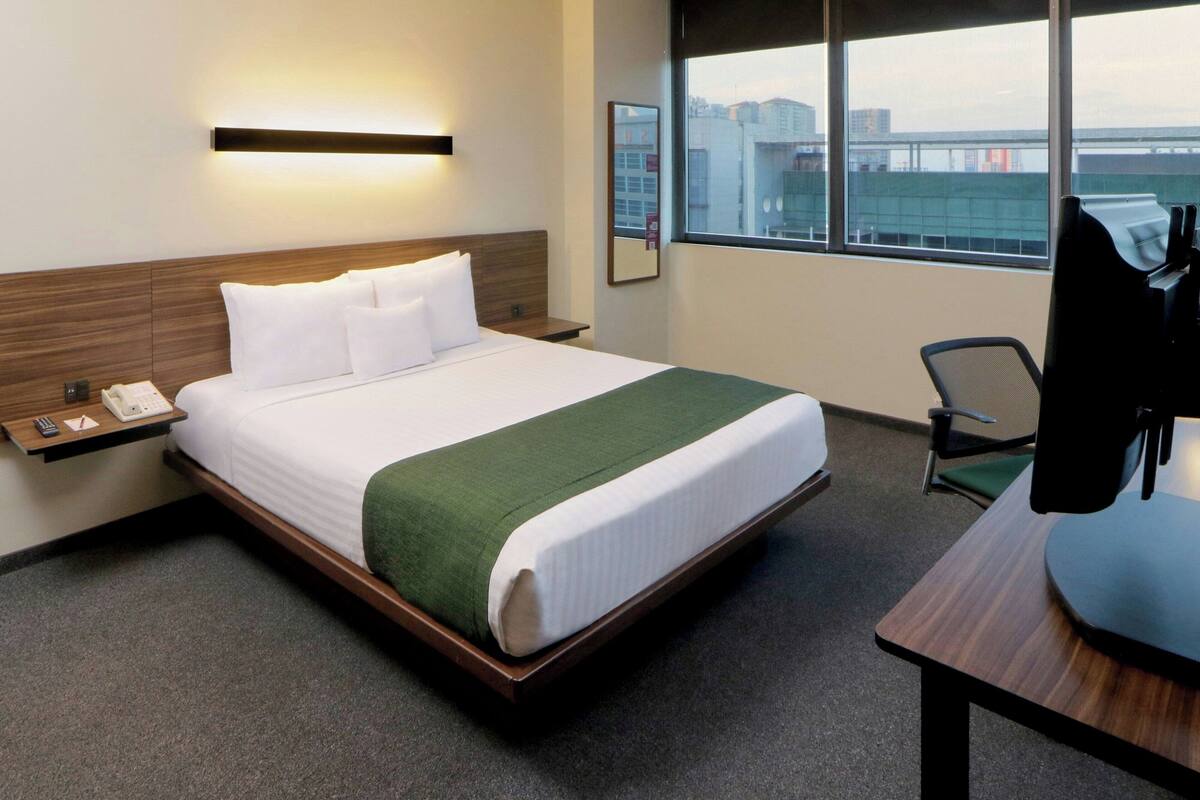 A hotel room with a bed, a desk, a chair, a phone, and a window with a city view.