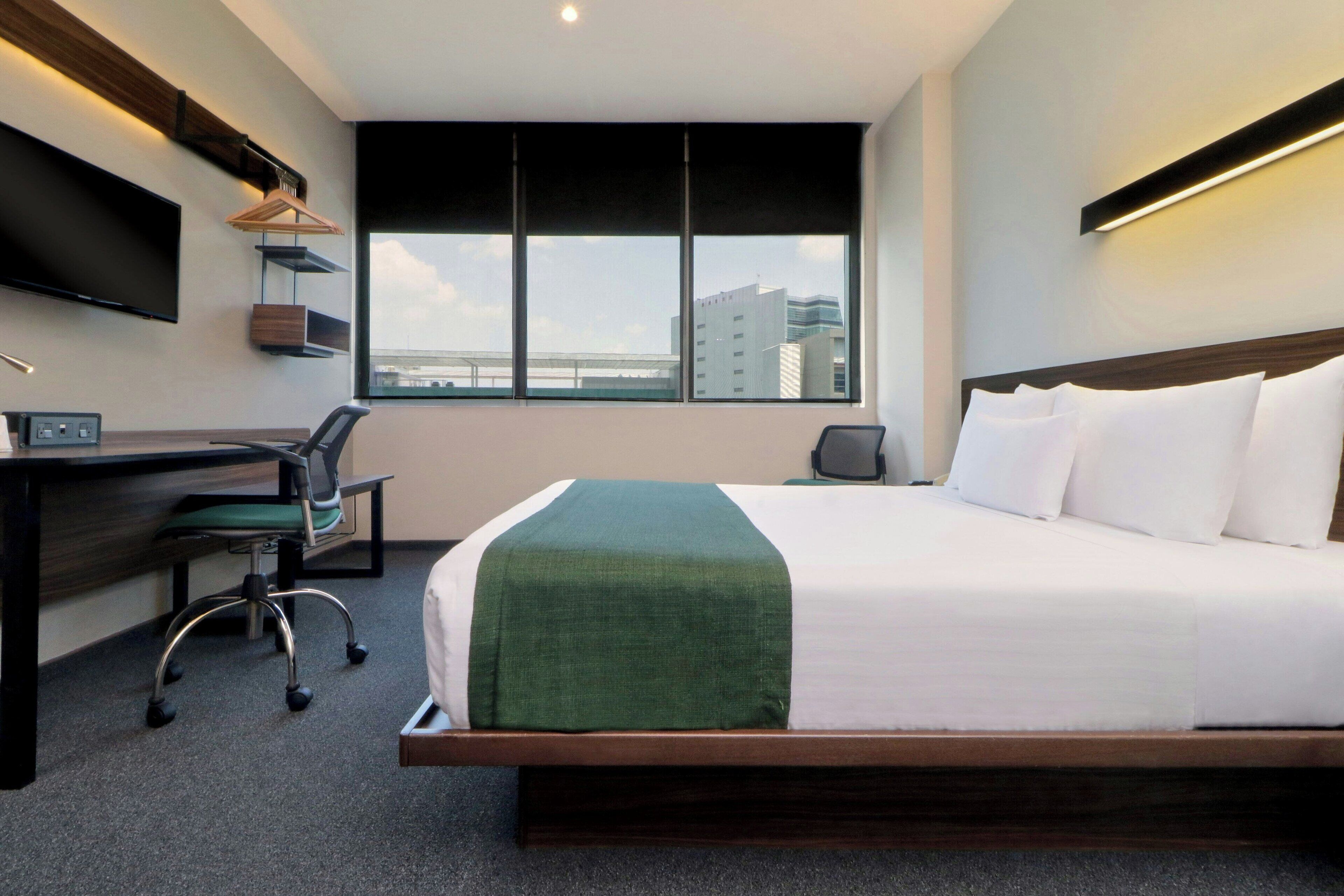 A hotel room with a large bed, a desk with a chair, a TV mounted on the wall, and a window with a view of buildings.
