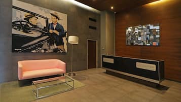 A reception area with a pink sofa, a glass coffee table, a painting of a car and two people, a modern desk, and a wall-mounted mirror.