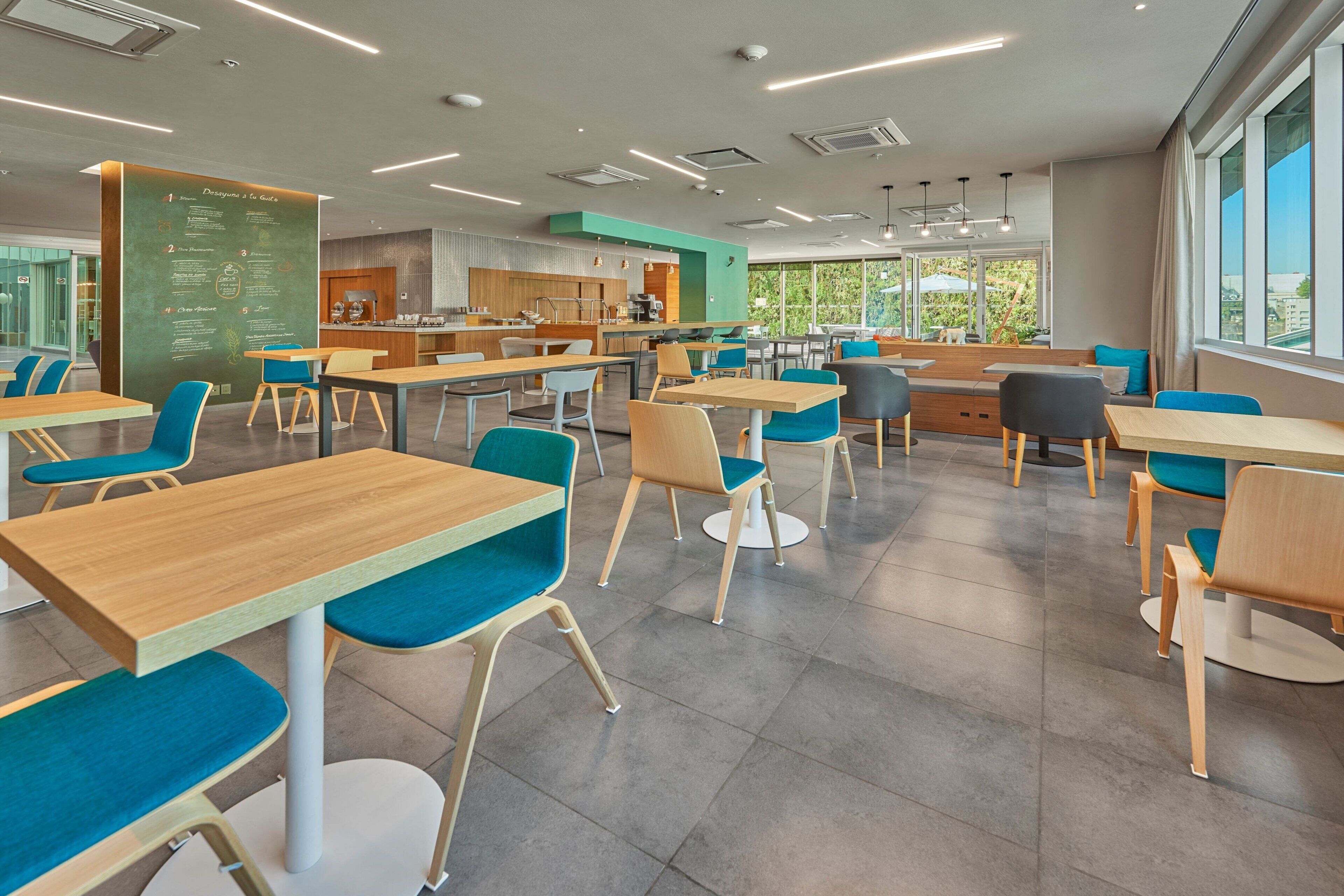 A modern dining area with wooden tables and blue upholstered chairs, a chalkboard menu, and large windows offering a view of the outdoors.
