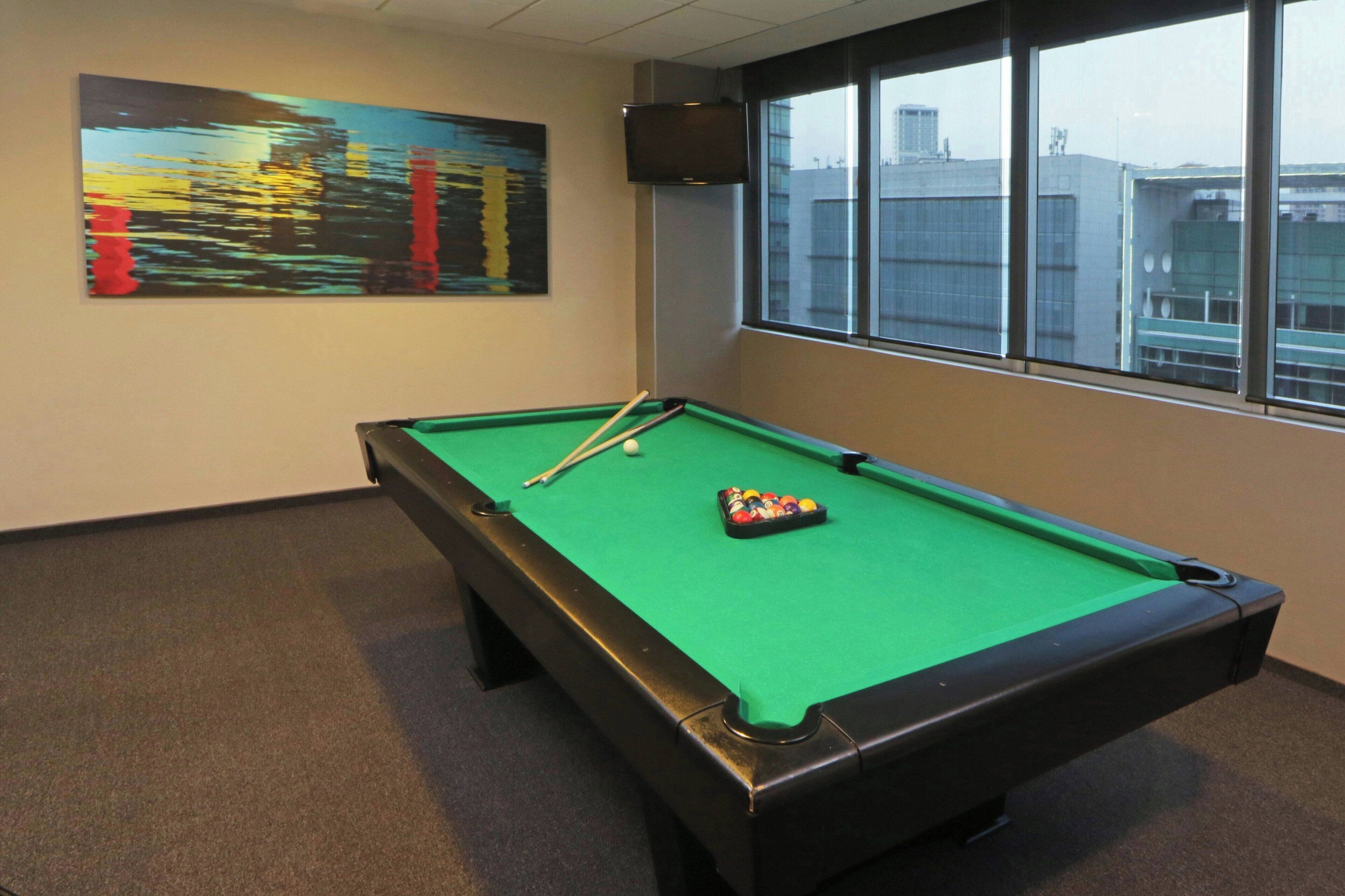A pool table in a room with a cityscape view through large windows, a painting on the wall, and a television mounted on the wall.