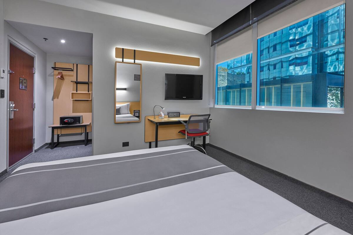 A modern hotel room with a bed, desk, chair, and a large window with a city view.