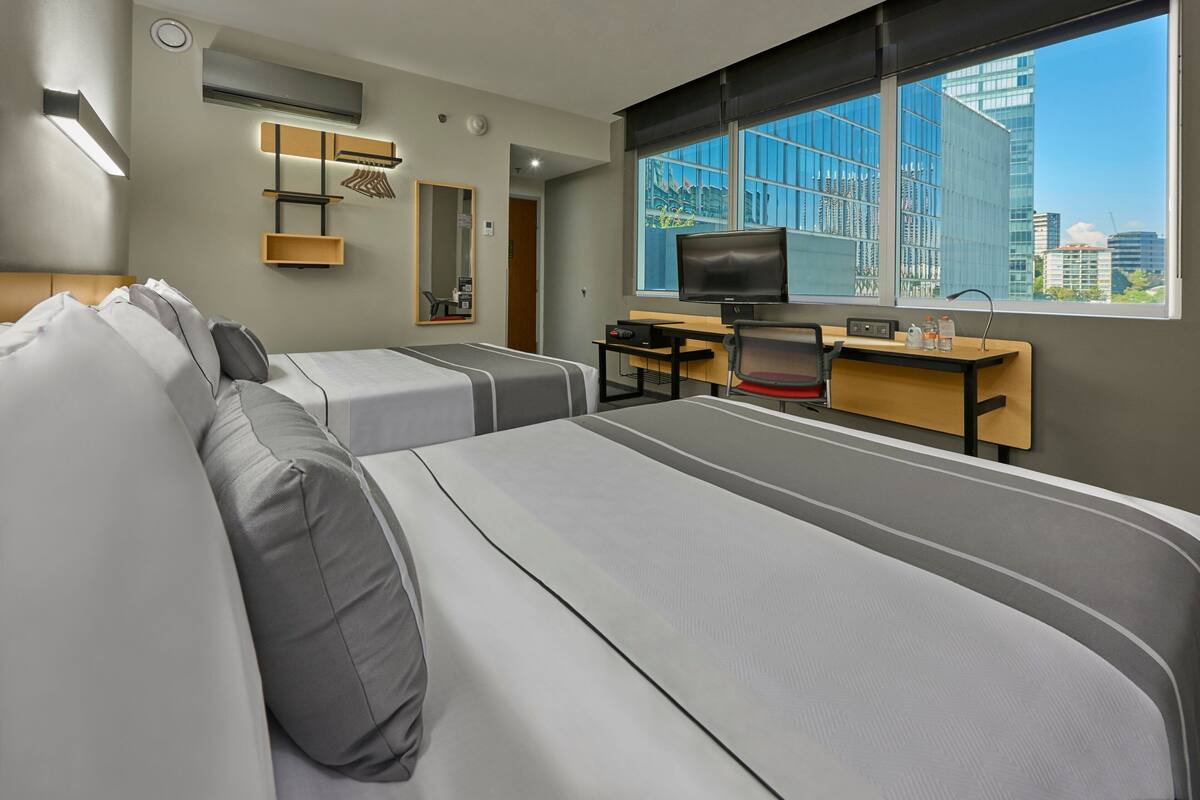 A modern hotel room with two beds, a desk with a computer, a TV, and a large window with a city view.