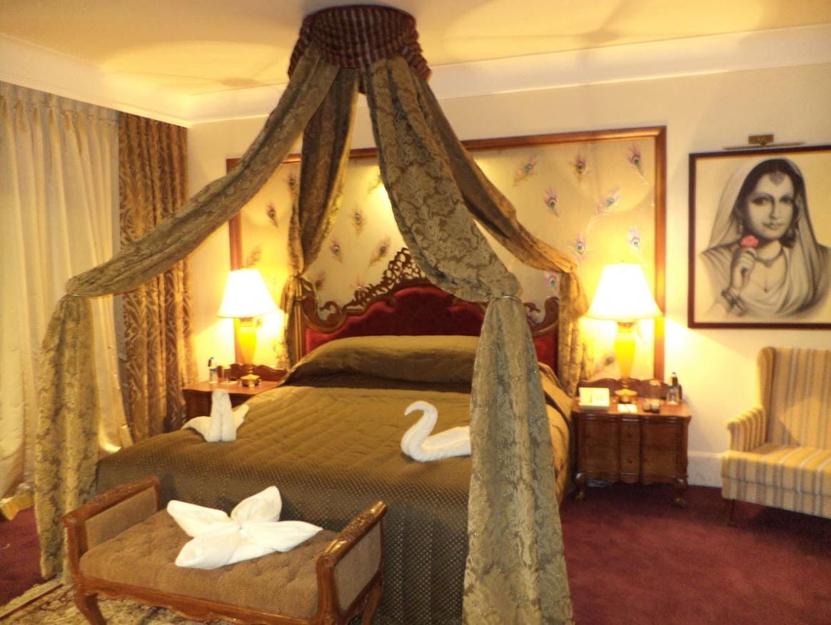 A hotel room with a four-poster bed, two bedside lamps, a chair, and a framed portrait on the wall.