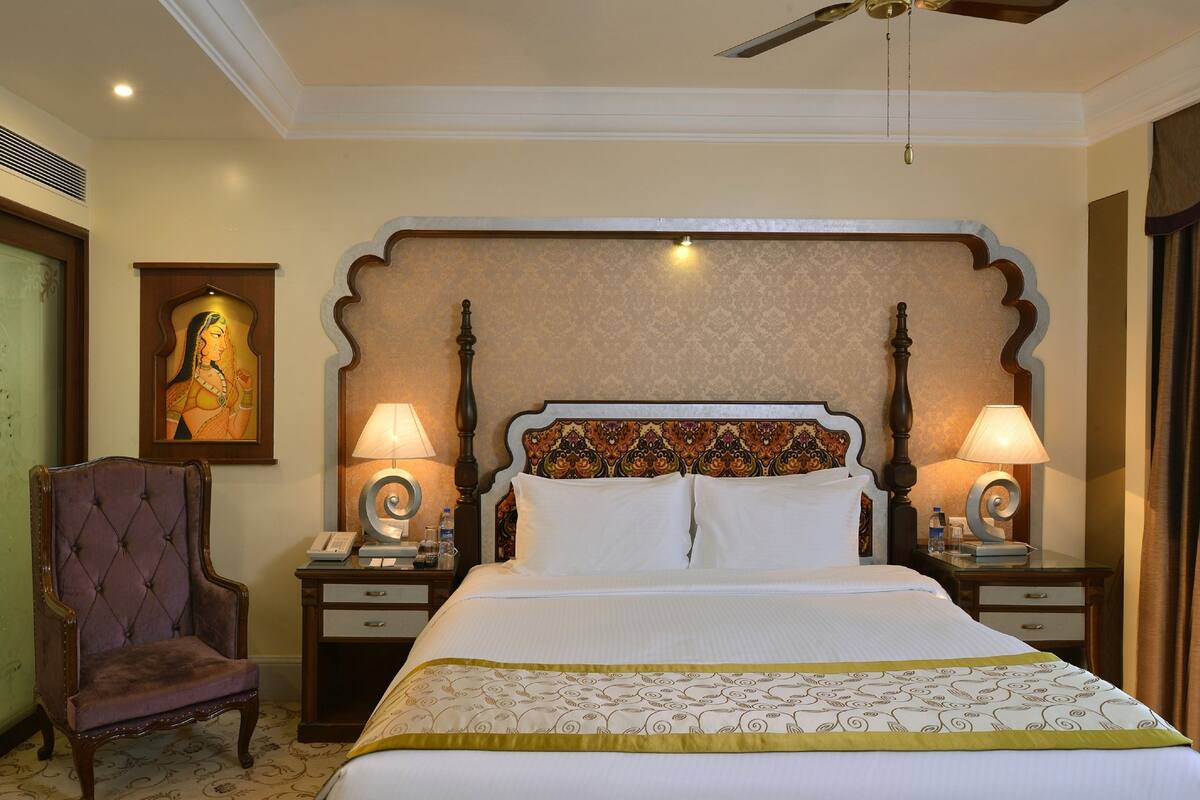 Een hotelkamer met een groot bed, twee nachtkastjes met lampen, een decoratief hoofdbord, een stoel en een schilderij aan de muur.