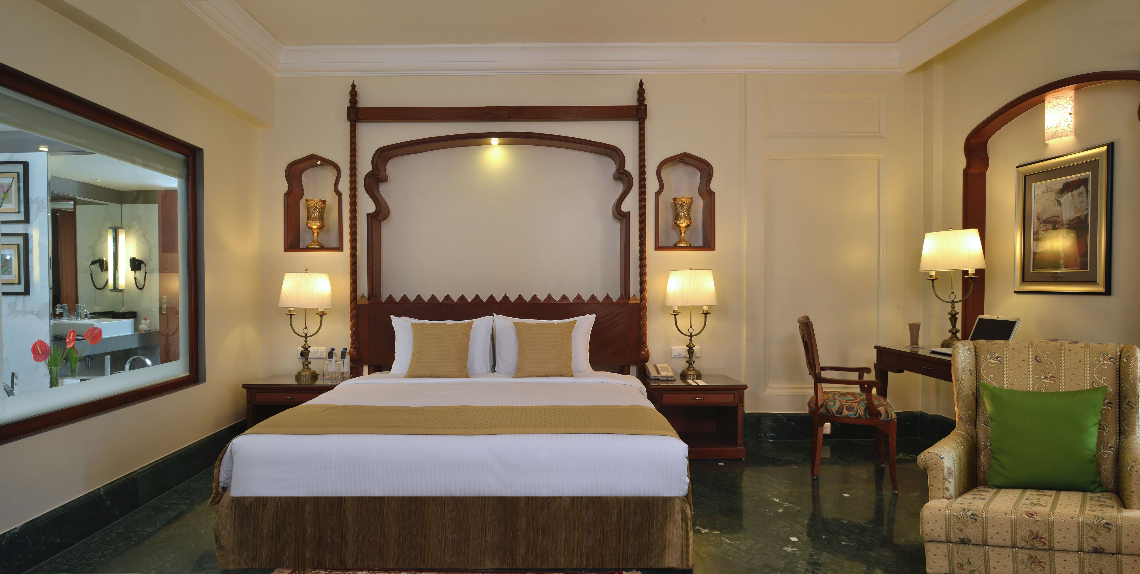 A hotel room with a large bed, two bedside tables with lamps, a chair with a green cushion, and a mirror on the wall.