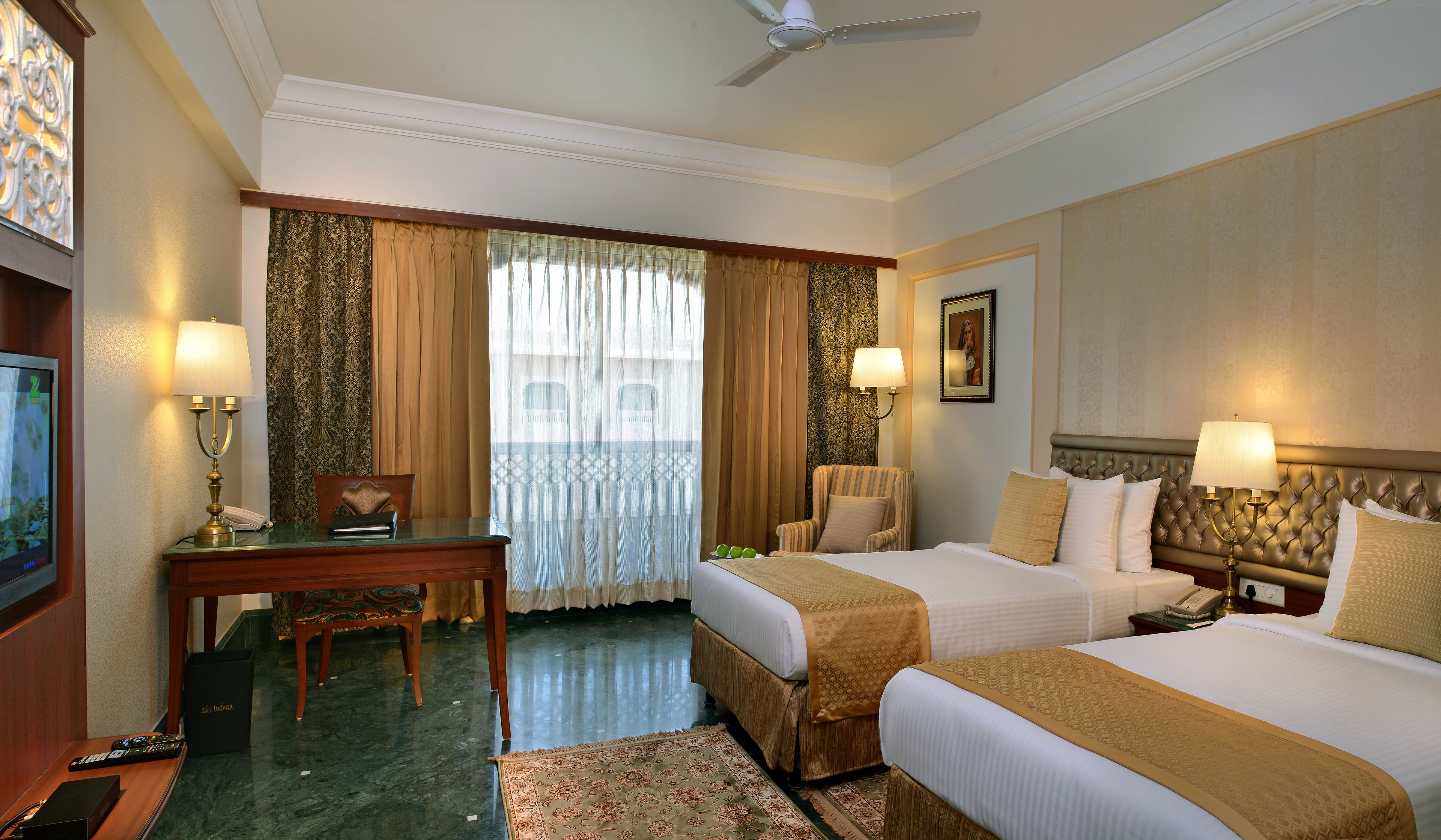 A hotel room with two beds, a desk, a chair, a TV, and a ceiling fan.