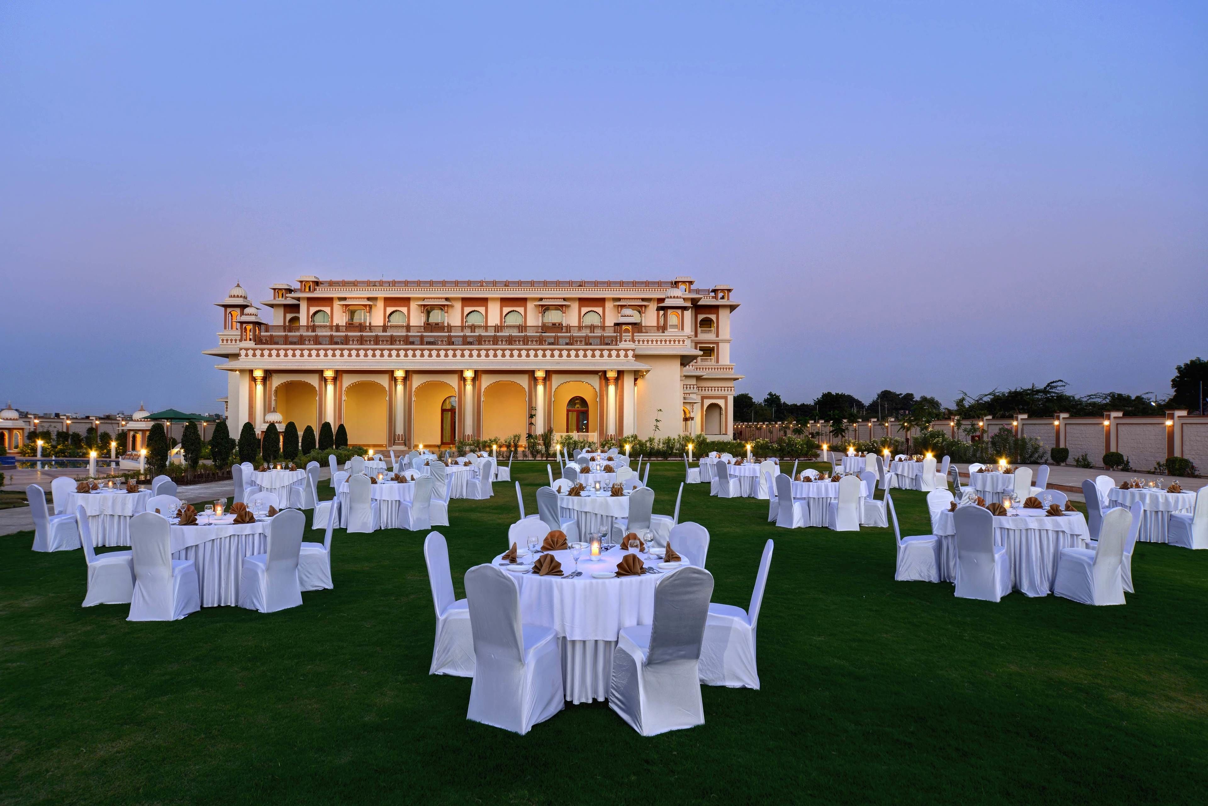 Un décor de banquet en plein air, avec des tables rondes et des chaises blanches, disposées sur une pelouse devant un grand bâtiment à l’architecture riche et élaborée.