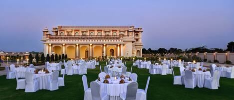 Un décor de banquet en plein air, avec des tables rondes et des chaises blanches, disposées sur une pelouse devant un grand bâtiment à l’architecture riche et élaborée.