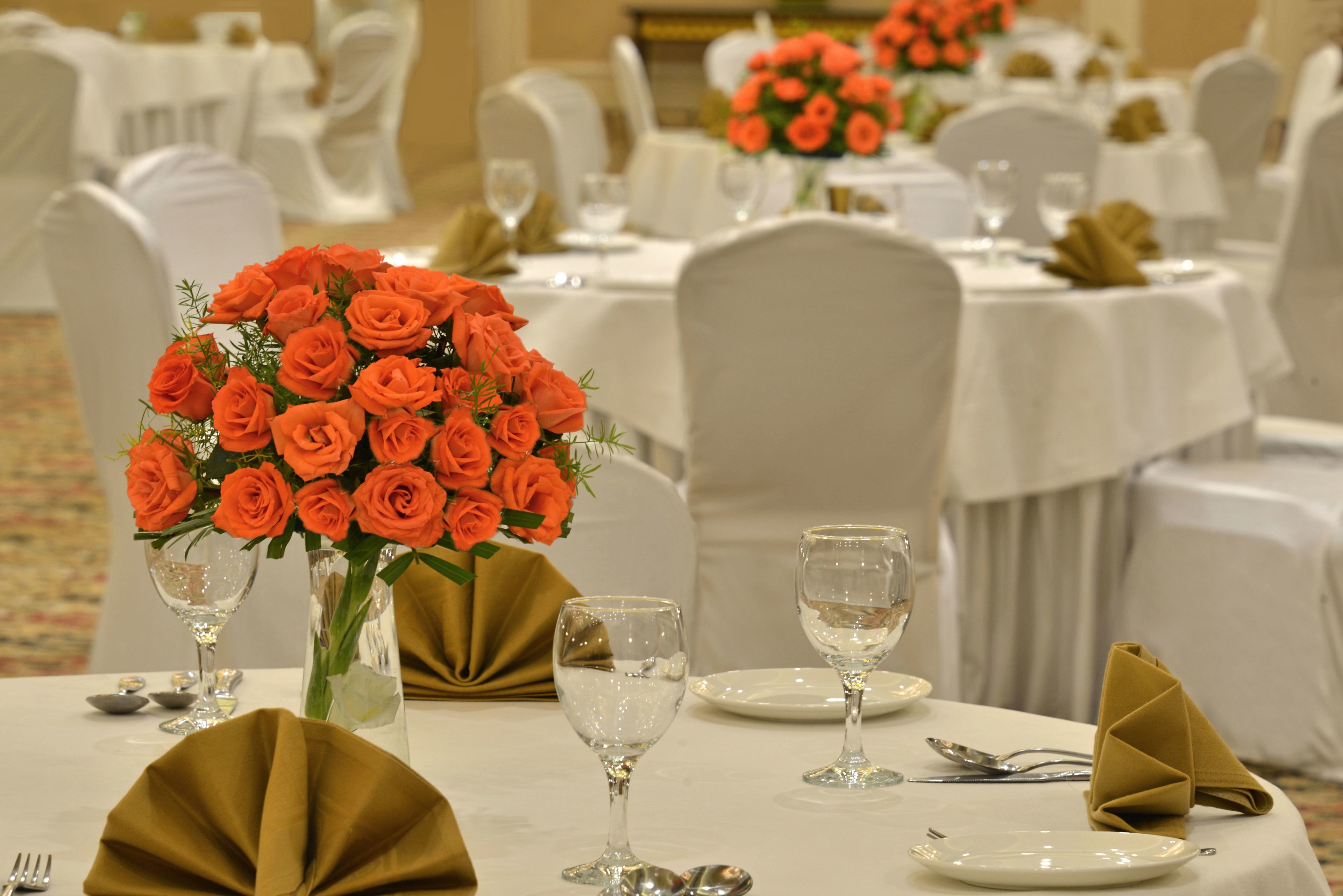 Un salón de banquetes con mesas redondas dispuestas para cenar, con manteles blancos, servilletas doradas y rosas anaranjadas en jarrones.