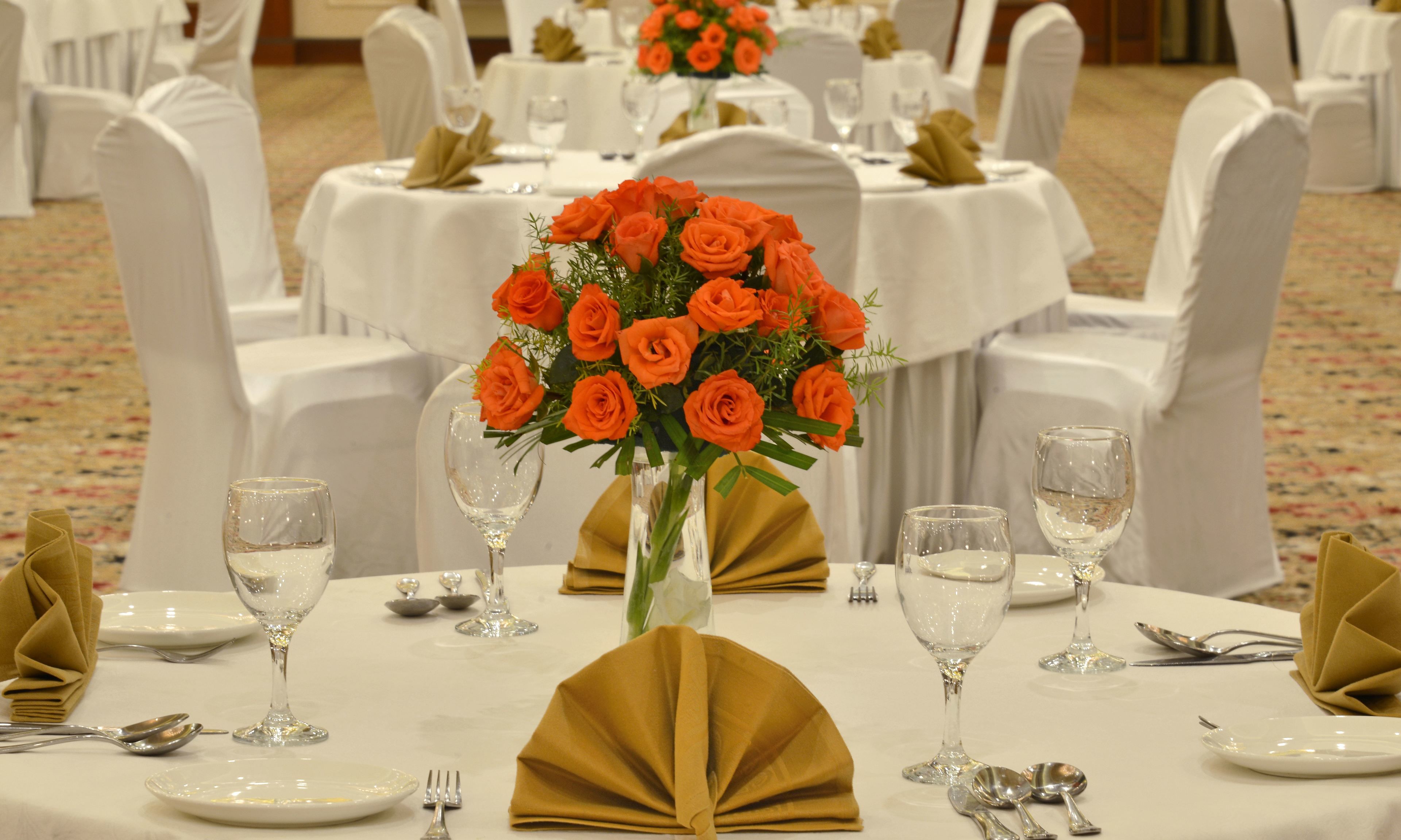 Ein festlich gedeckter Tisch mit weißen Tischdecken, goldenen Servietten und einem Strauß aus orangefarbenen Rosen in einer klaren Vase.