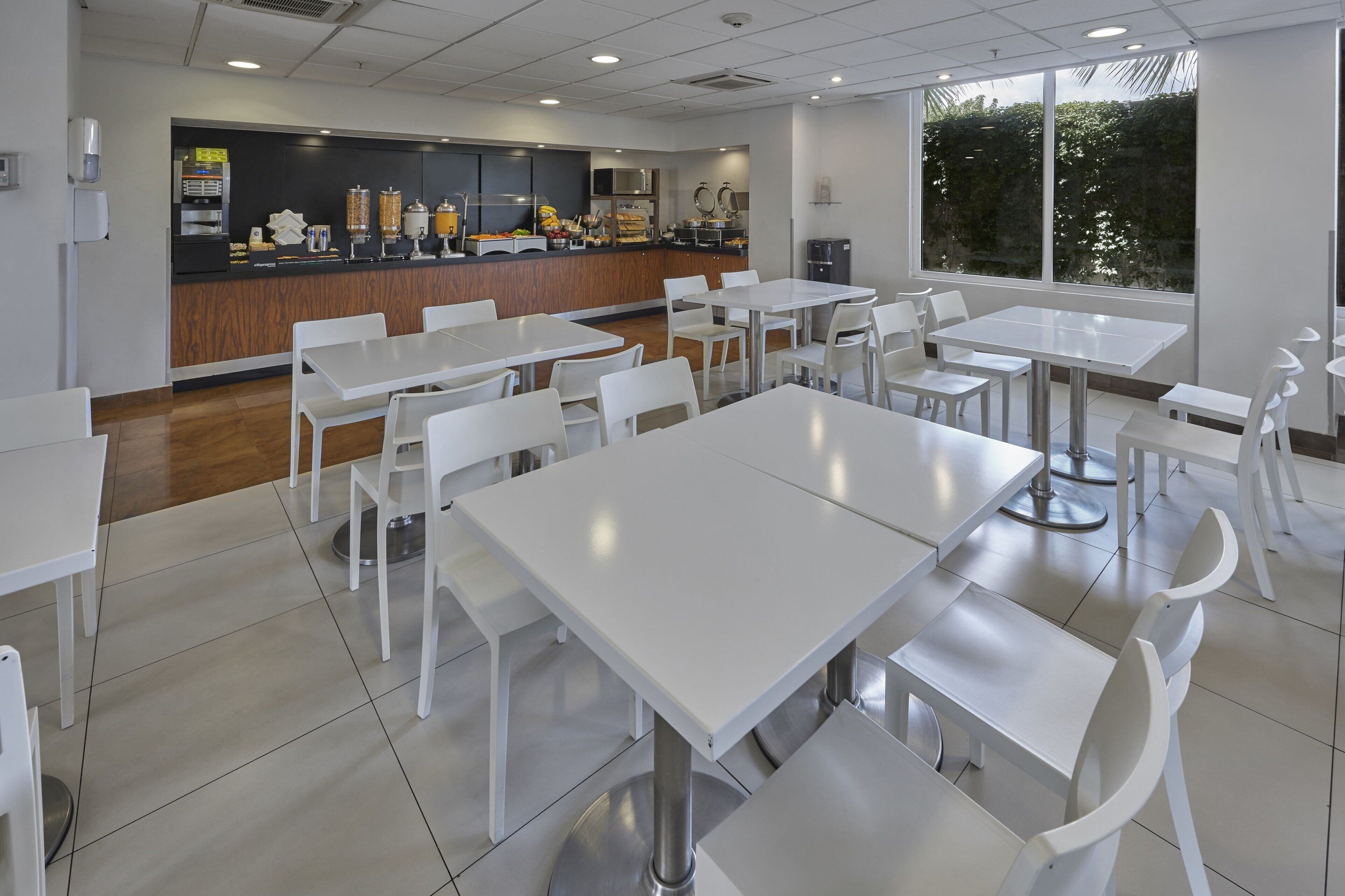 A modern dining area with white tables and chairs, a counter with food items, and large windows providing natural light.